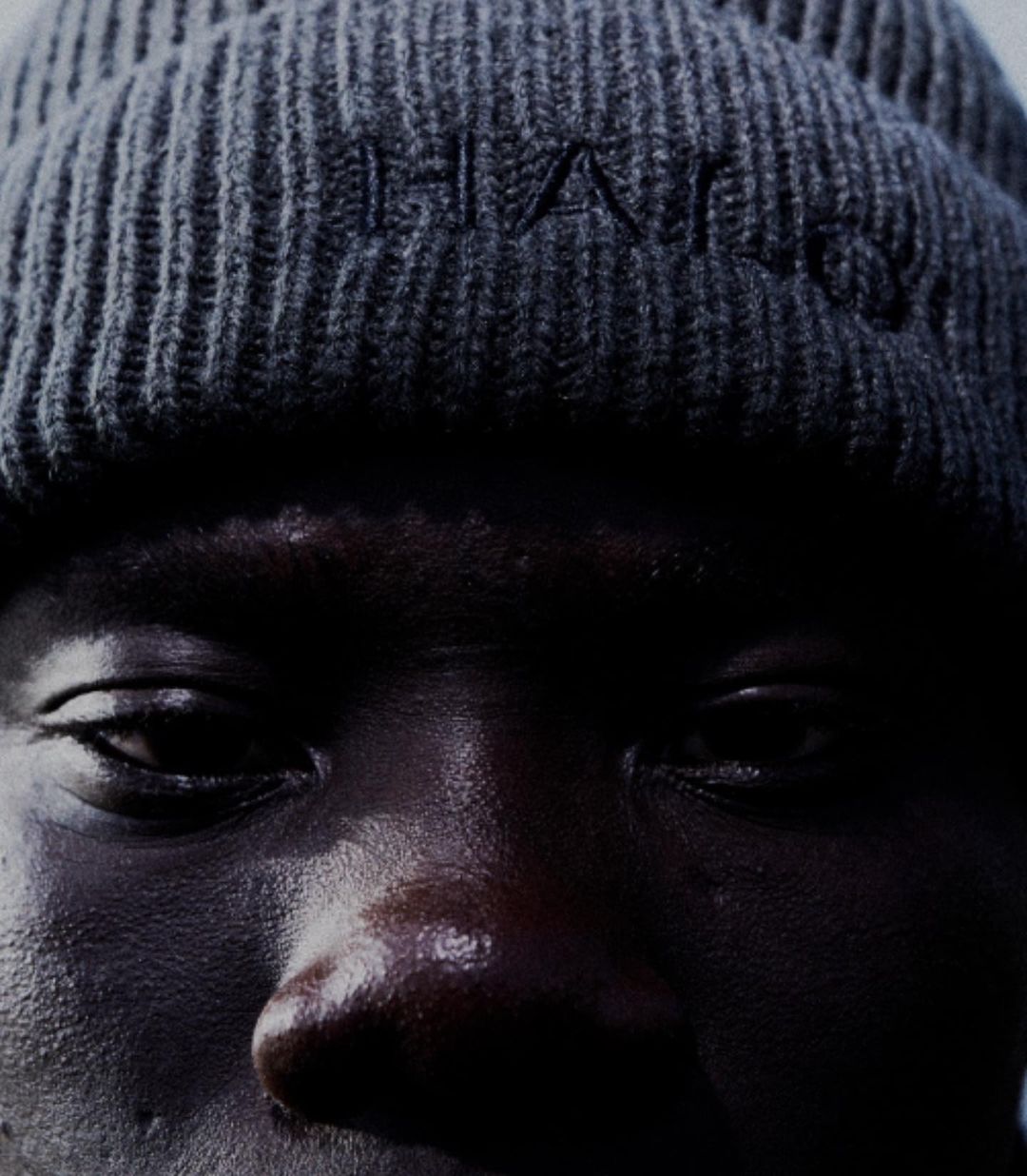 Close-up portrait of an adult of African descent wearing a gray knit beanie with embroidered letters under dark, shadowy lighting.