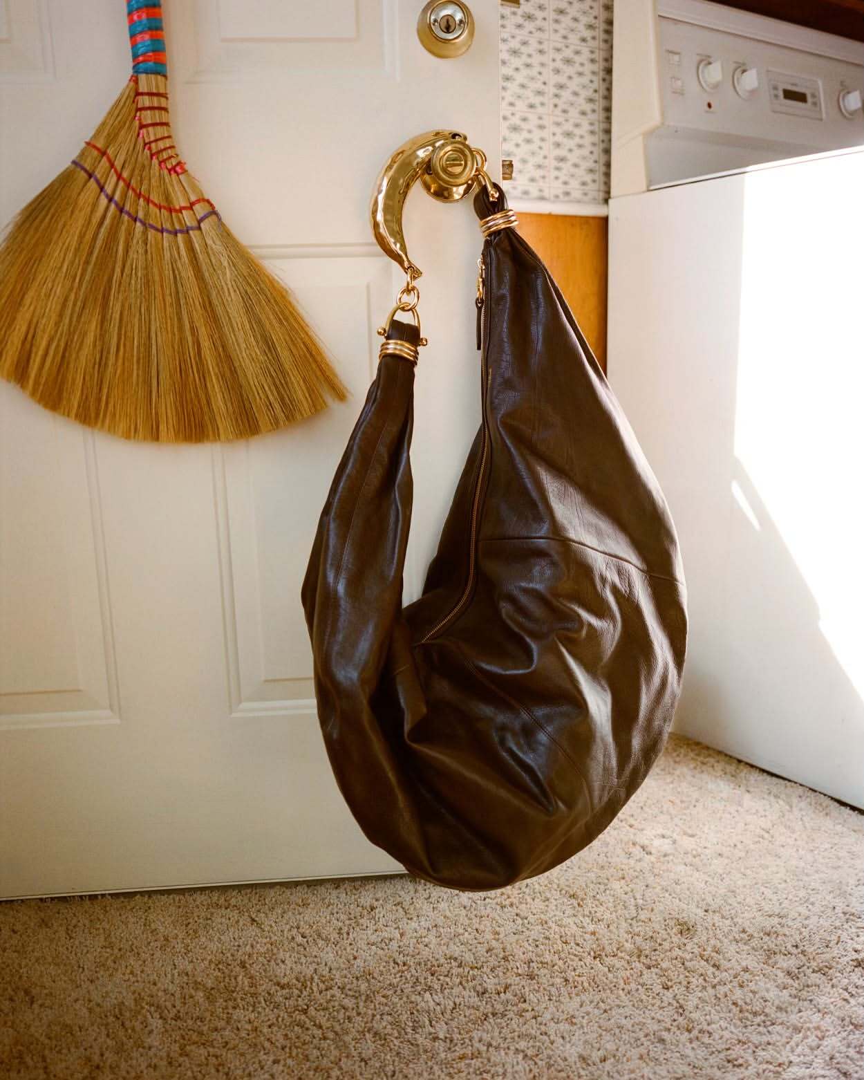 Dark brown leather hobo bag hangs from ornate door handle, set against a white door, and a broom in a domestic interior.