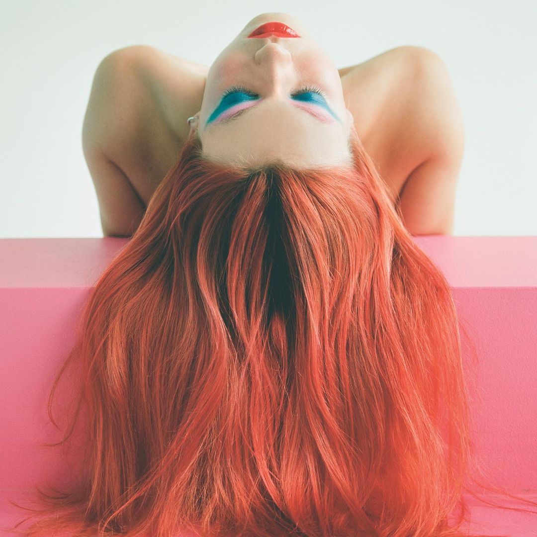 Woman with red hair and blue eye makeup leans on a pink box in a beauty shot against a white background in a studio setting.
