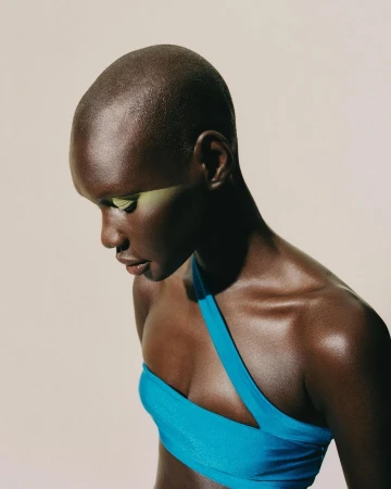 Close-up portrait of a bald Black woman wearing a bright blue top with striking green eyeshadow against a light background