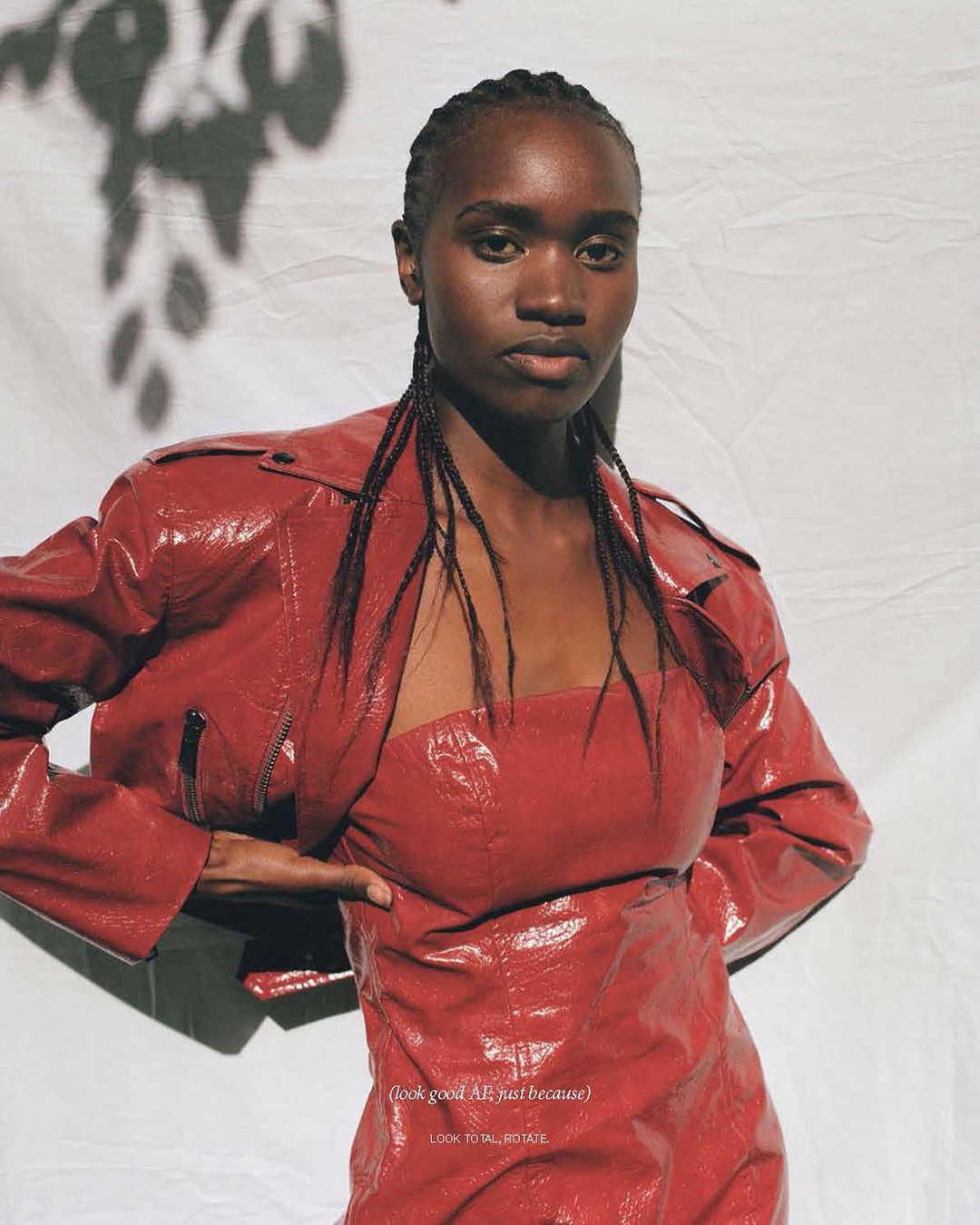 Woman in a red patent-leather dress and jacket stands indoors against a white background with shadows, modelling the garment for fashion.