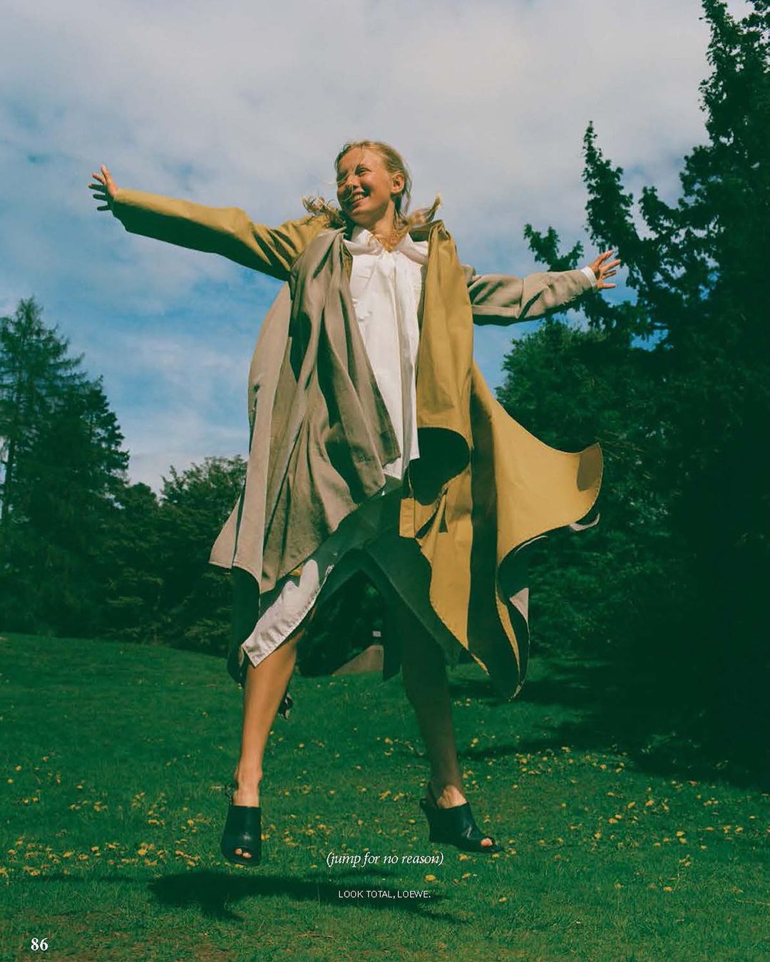 Woman in a tan coat jumping in the air with arms outstretched in a grassy park under a partly cloudy sky, fashion editorial.