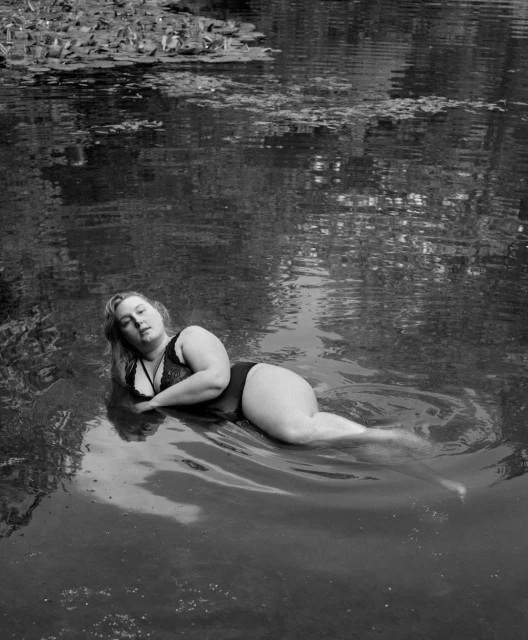 Adult woman with blond hair lies in dark water with lily pads in the background in a black and white, eye-level photograph.