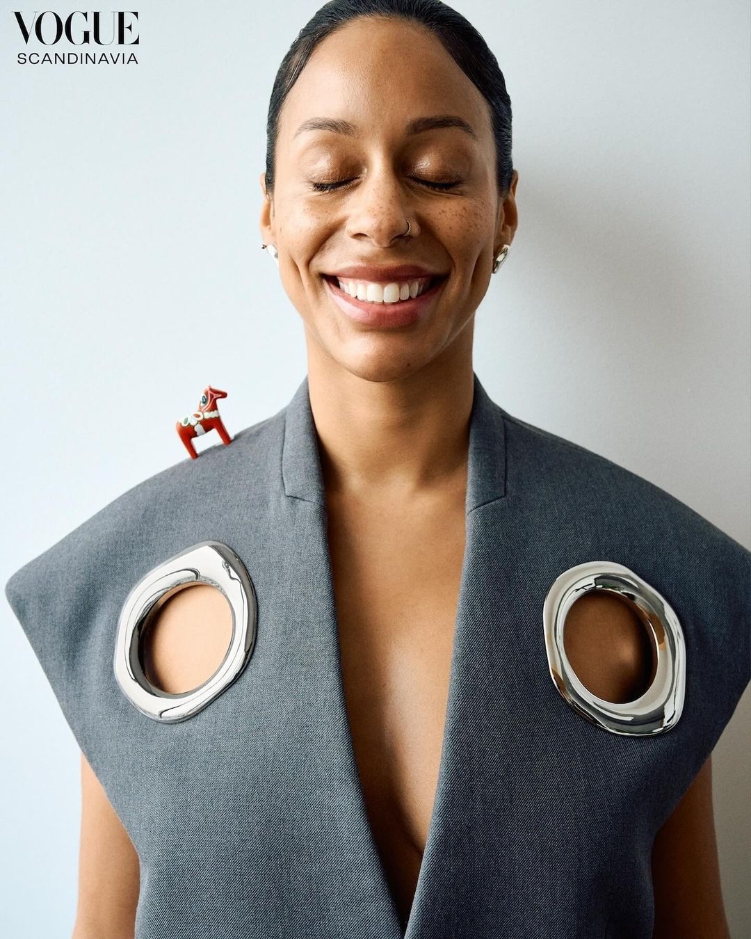 Woman with a grey sleeveless jacket smiles with her eyes closed, featuring a red toy horse on her shoulder and Vogue Scandinavia logo.