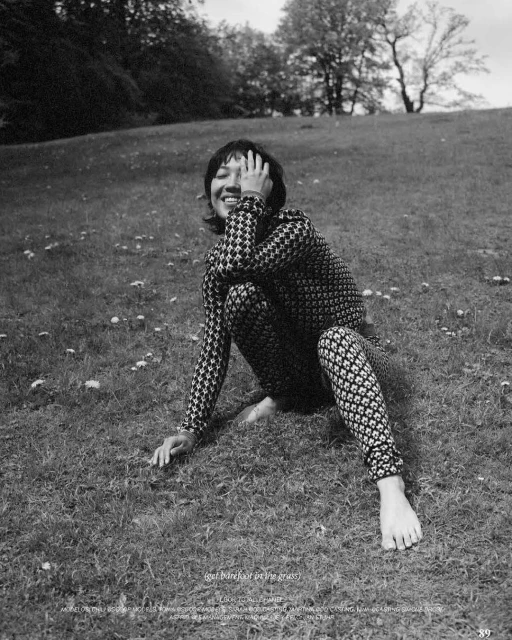 Smiling woman with patterned clothing seated in grass. Outdoor fashion portrait with black-and-white photography. Candid, relaxed posture and natural setting.