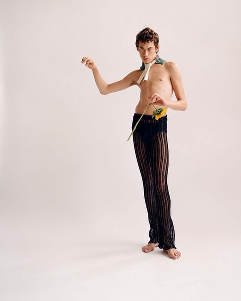 Nude male fashion model poses with sunflower against white background. He wears sheer black pants and neck scarf for a minimalist style portrait.