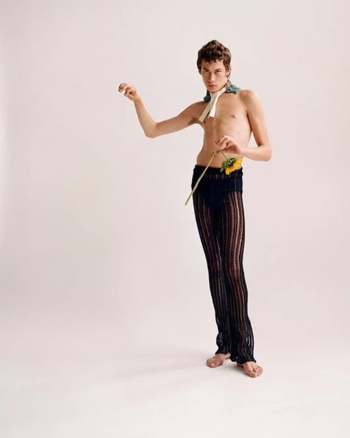 Nude male fashion model poses with sunflower against white background. He wears sheer black pants and neck scarf for a minimalist style portrait.