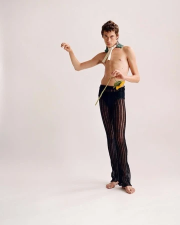 Nude male fashion model poses with sunflower against white background. He wears sheer black pants and neck scarf for a minimalist style portrait.