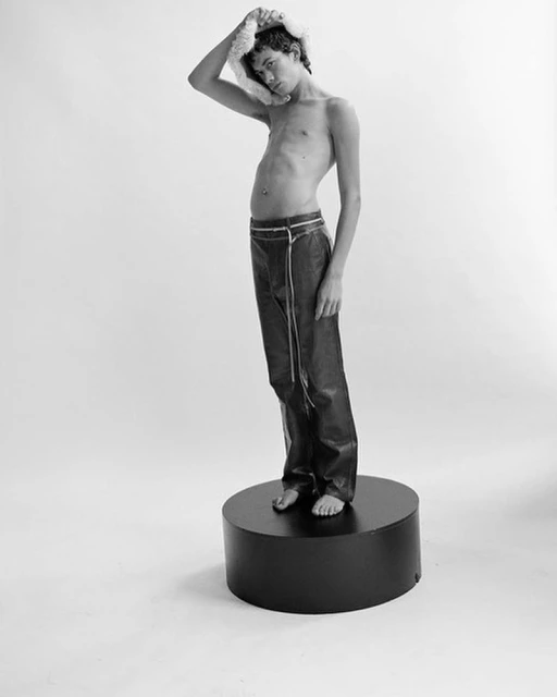 Monochrome studio shot of a young adult man standing barefoot on a black platform, wearing dark pants, bare-chested, holding a fluffy hat.