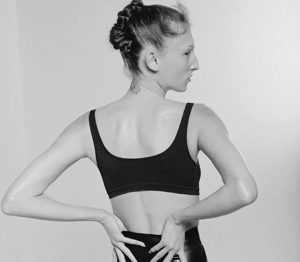 Black and white portrait of a woman with a braided updo wearing a sports bra with her hands on her lower back in a studio setting.