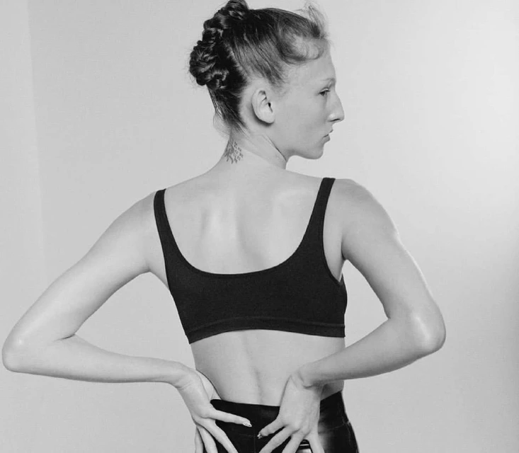 Black and white portrait of a woman with a braided updo wearing a sports bra with her hands on her lower back in a studio setting.