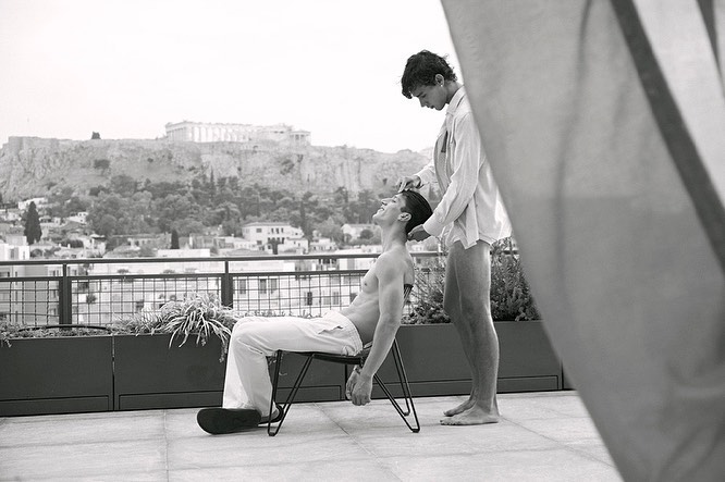 Black and white photograph of two young adult men on a rooftop, one seated as the other stands behind him, with city skyline backdrop.