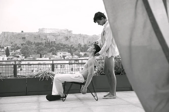 Black and white photograph of two young adult men on a rooftop, one seated as the other stands behind him, with city skyline backdrop.