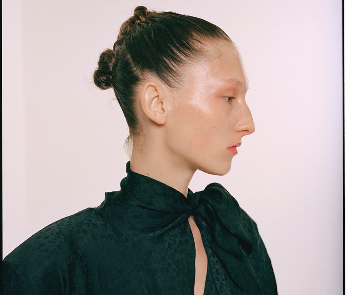 Profile of a woman with dark hair styled in braided knots, light skin, dark top with bow, in a beauty or fashion context.