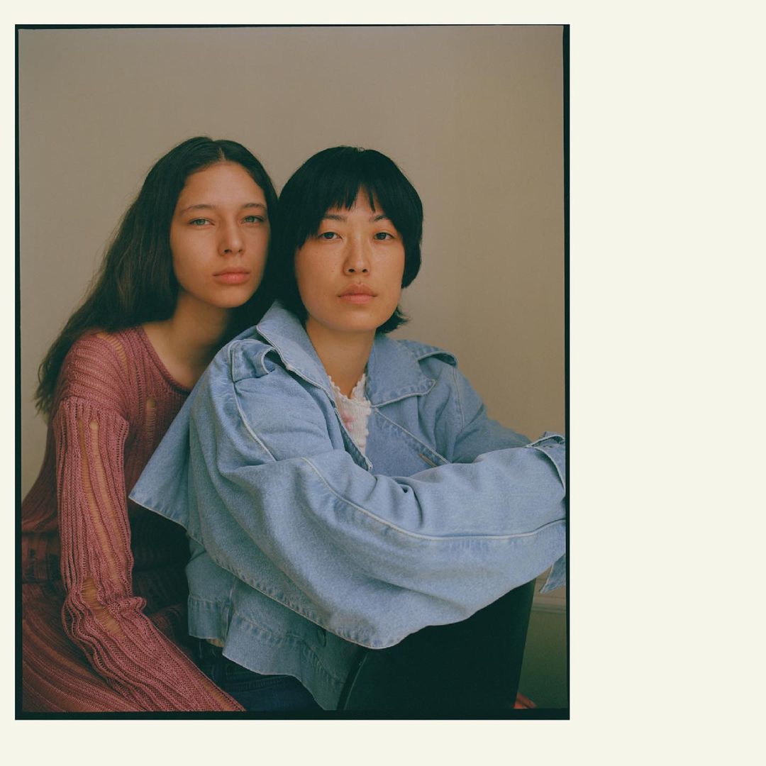 Two young women pose indoors: one in a denim jacket and black pants, the other in a long-sleeved pink top, looking directly at the camera.