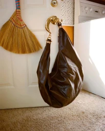 Dark brown leather hobo bag hanging from a gold doorknob on a white door inside a domestic room by Dennis Stenild