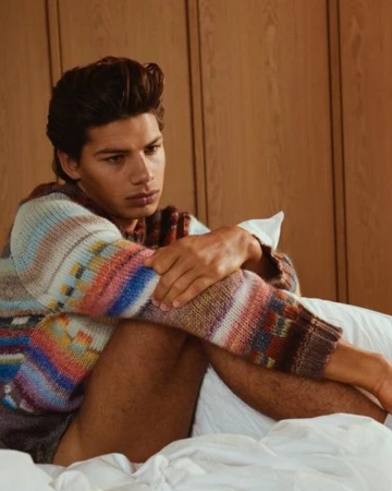 Young man in a multi-colored patterned knit sweater seated on a white bed with wooden paneling behind him.