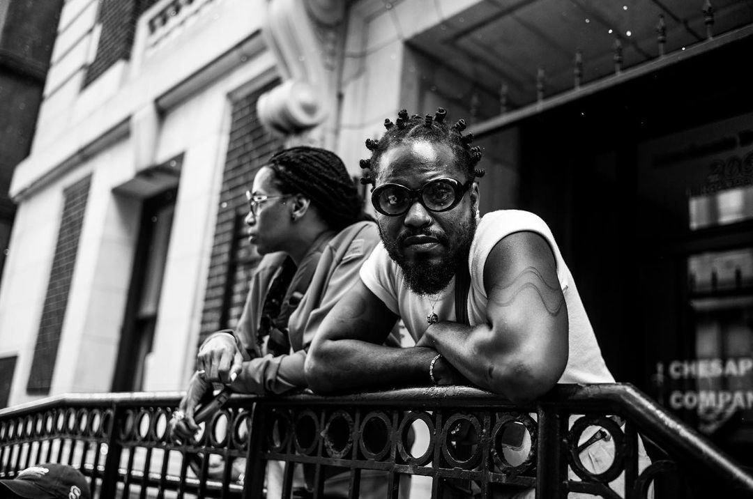 Black and white portrait of two adult humans, one with knotted hair and sunglasses, posing in front of a building on a city street.