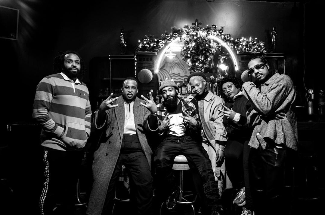 Black and white shot of six people posing together in a bar setting, festive decorations hanging in background.
