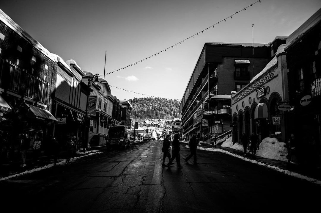 Black and white street scene with people crossing the road in a town lined with buildings, under a sky with distant, snow-covered trees.