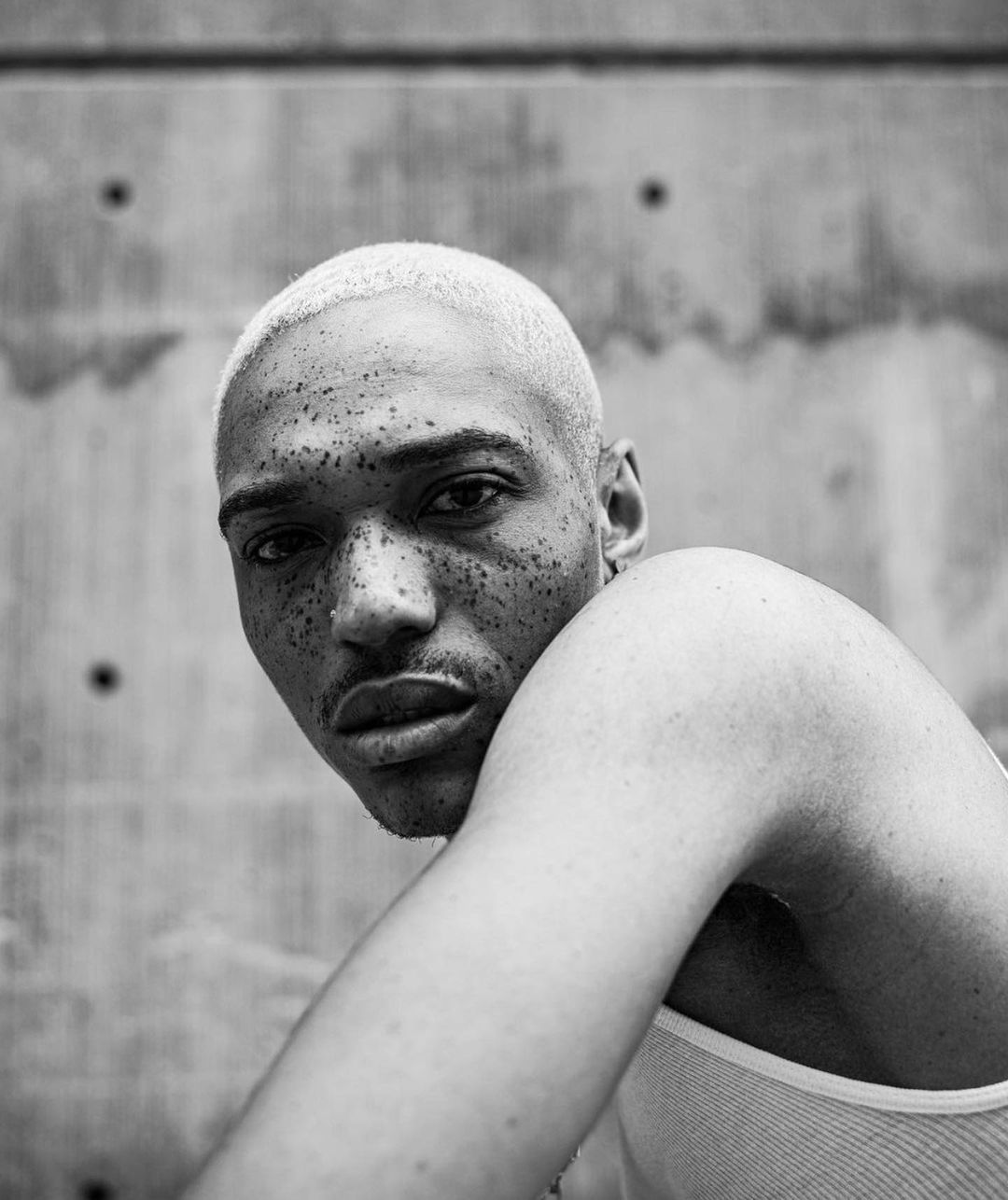 Black and white portrait of a young man with short blonde hair and freckles posing against a textured grey wall, close-up studio shot.