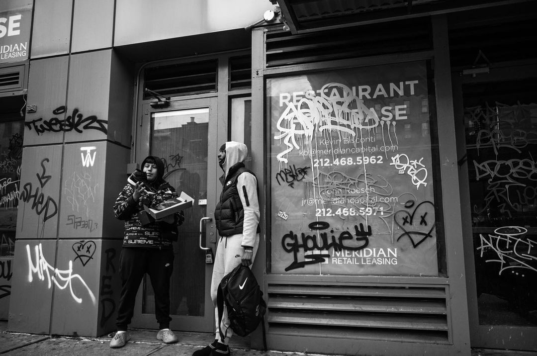 Two men stand outside a building covered in graffiti. One holds a shoebox and the other has a backpack in an urban scene.