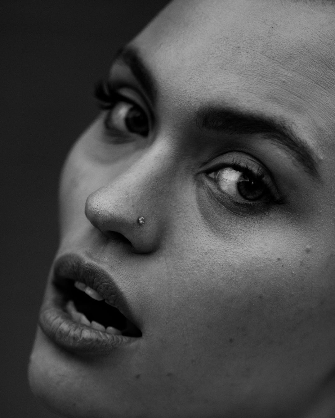 Close-up black and white portrait of an adult woman with fair skin, dark eyes, and a nose piercing looking upwards.