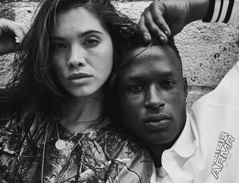 Black and white portrait of a young interracial couple, the woman in a patterned top, the man in a white shirt with the Under Armour logo.