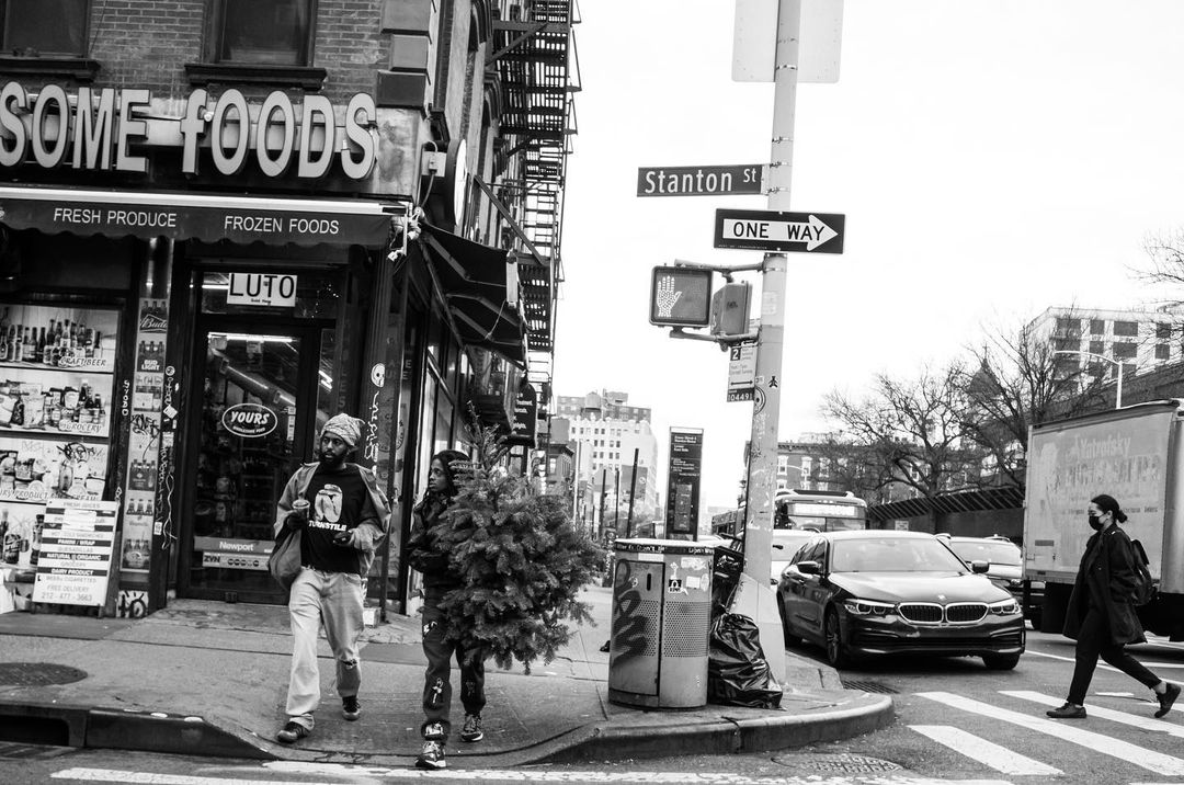 Black and white street scene with two people carrying a Christmas tree, a woman crossing the street, and a store with fresh produce.