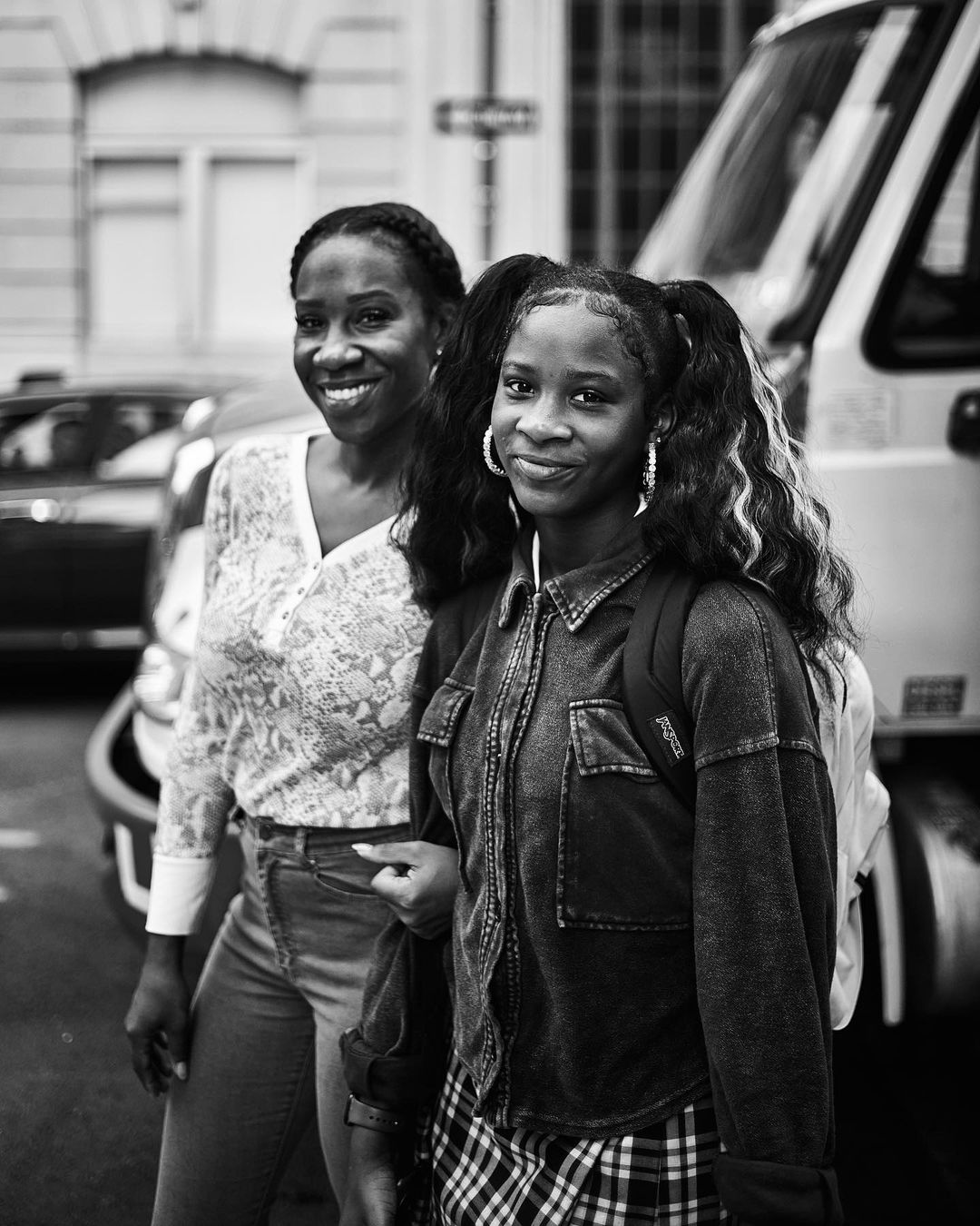 Black and white portrait of an adult and a child standing on a city street in front of a large vehicle, both smiling at the camera.