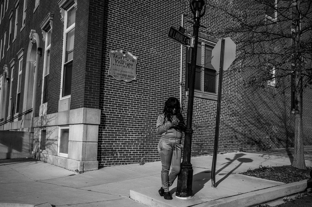 Monochrome shot of a young woman leaning against a lamppost using her cellphone in an urban setting with a brick building.