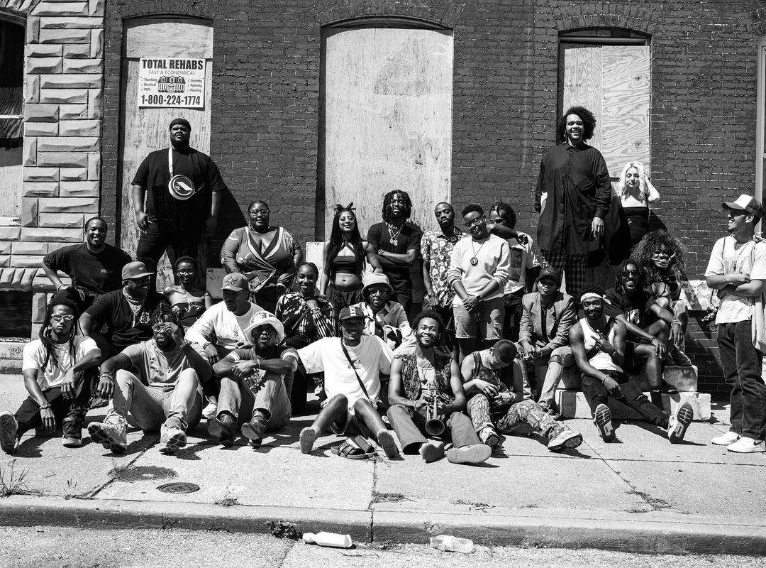 Large group of diverse adults and young adults gathered outdoors on a sidewalk in front of a brick building for a group portrait.