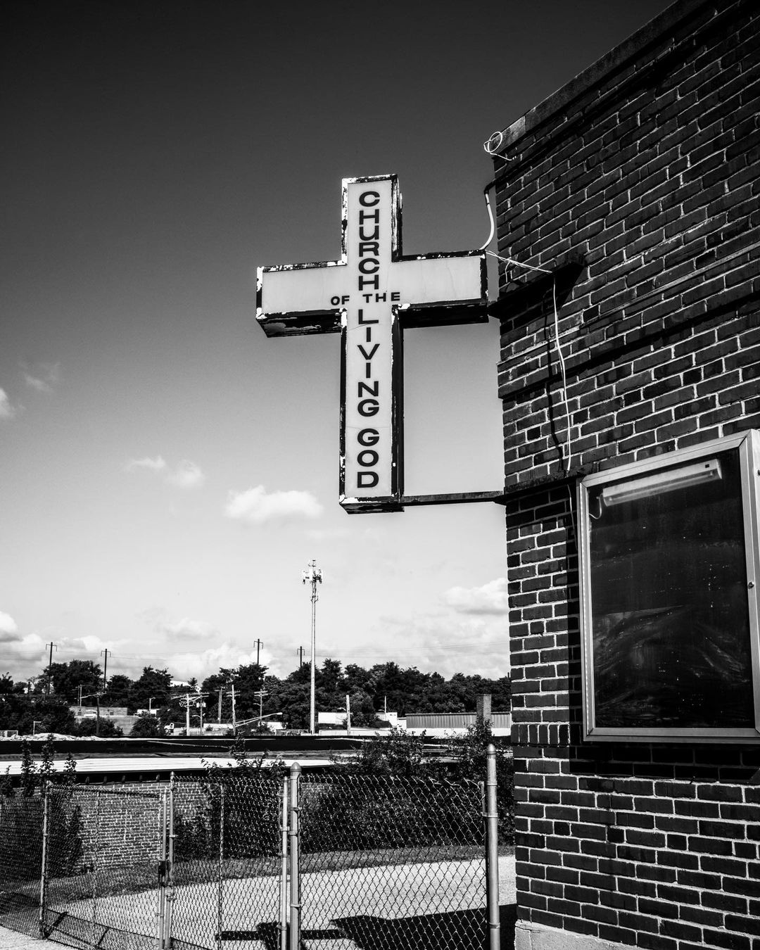 Black and white photograph of a brick building with a 'Church of the Living God' sign in the shape of a cross.