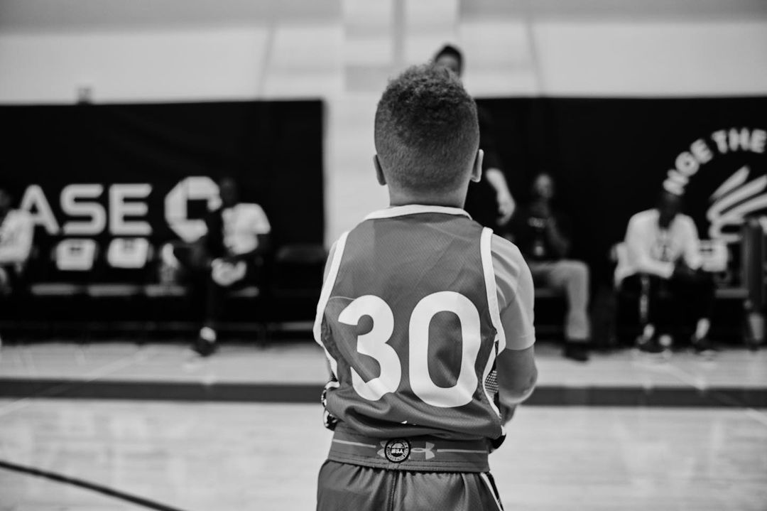 Boy in basketball jersey with number 30 stands on basketball court, facing away from camera with people blurred in background.