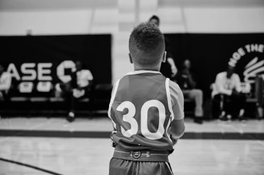 A young child wearing a basketball jersey with the number 30 stands on an indoor court facing away from the camera.