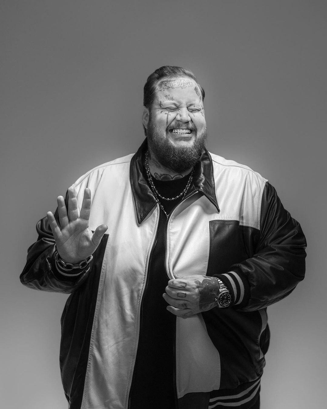 Monochromatic portrait of a smiling, bearded man with face tattoos wearing a jacket, chain necklace and wristwatch against grey backdrop.