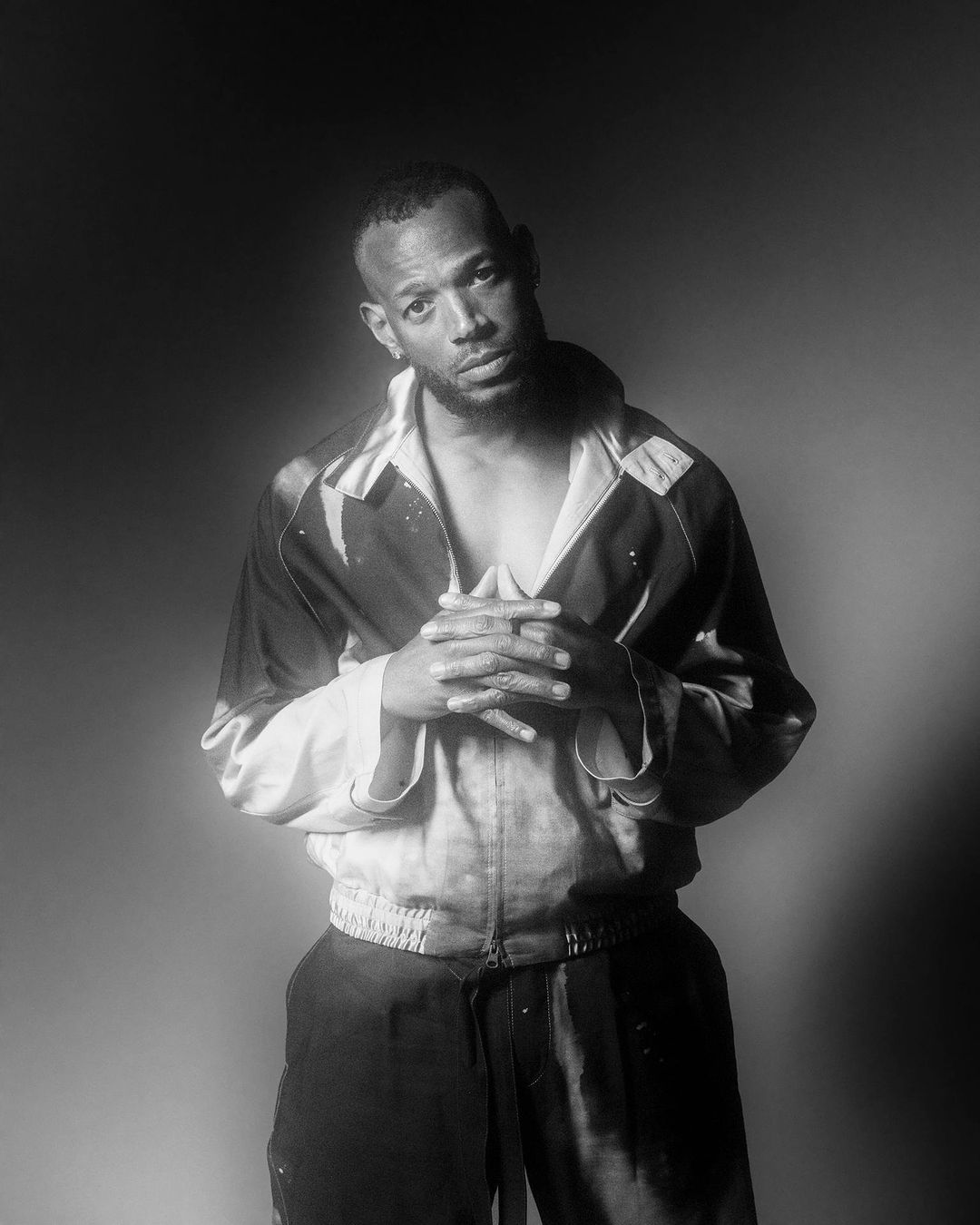 Black-and-white portrait of a man wearing a patterned jacket with clasped hands against a gradient studio backdrop.
