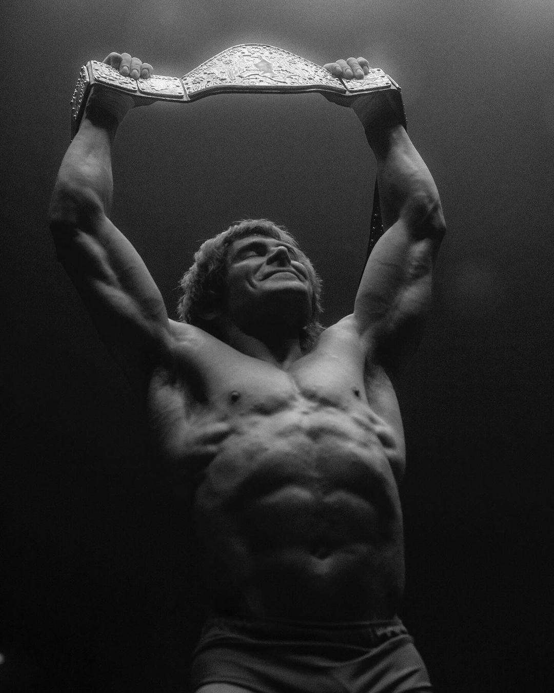 Monochromatic shot of a muscular adult male athlete holding a championship belt overhead in a dark indoor environment under bright lighting.