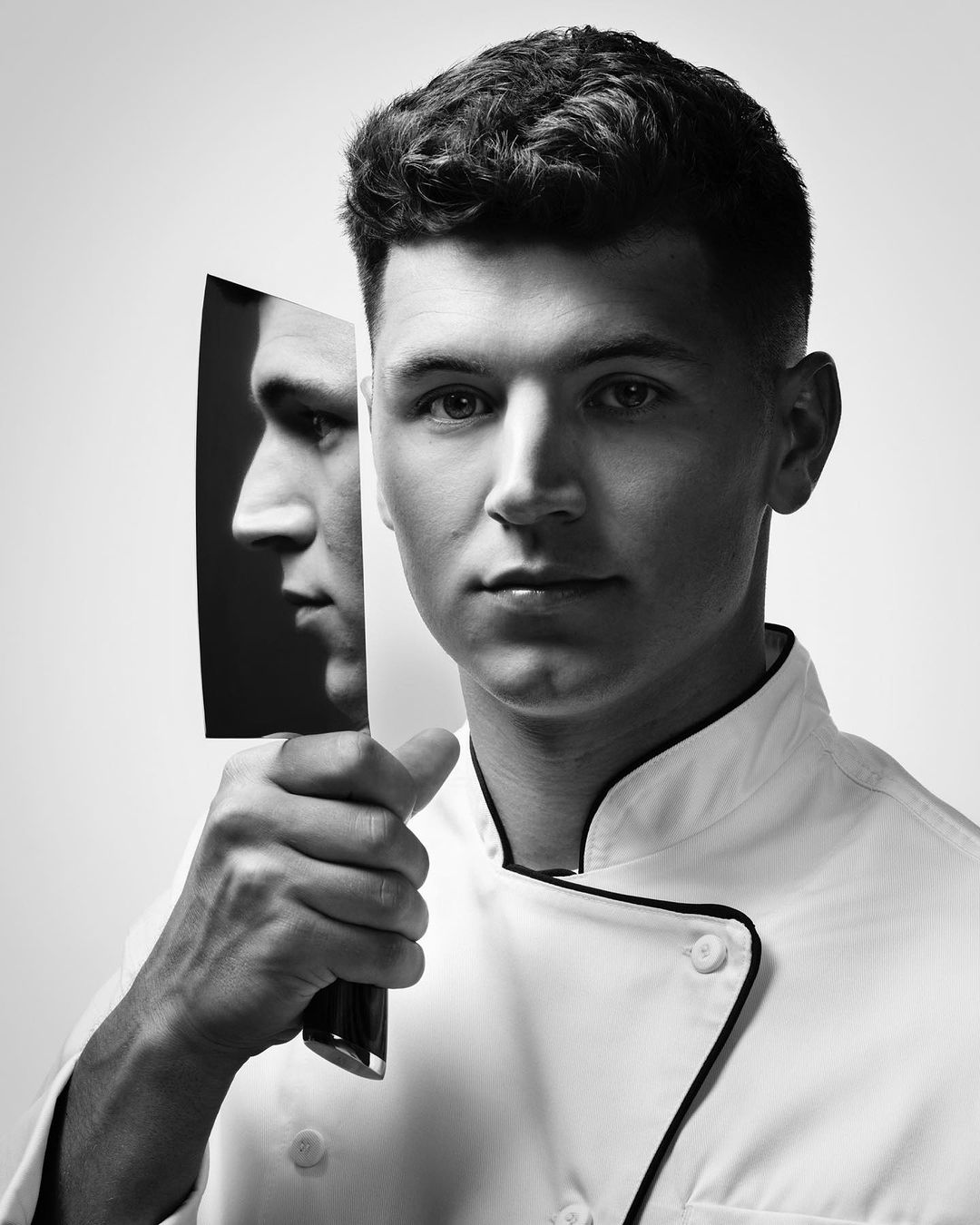 Black and white portrait of a young man in a chef's coat holding a knife that reflects his face in profile against a neutral background.