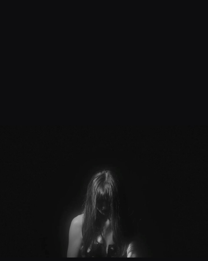 Monochromatic studio portrait of a female with long dark hair partially covering her face, wearing a dark strappy top against a black background.