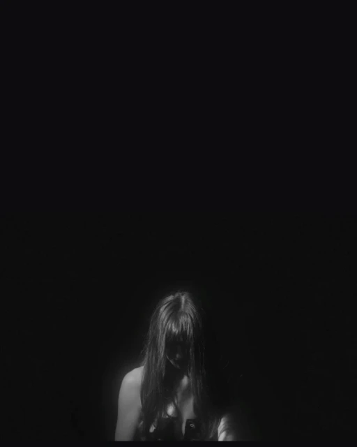 Monochromatic studio portrait of a female with long dark hair partially covering her face, wearing a dark strappy top against a black background.