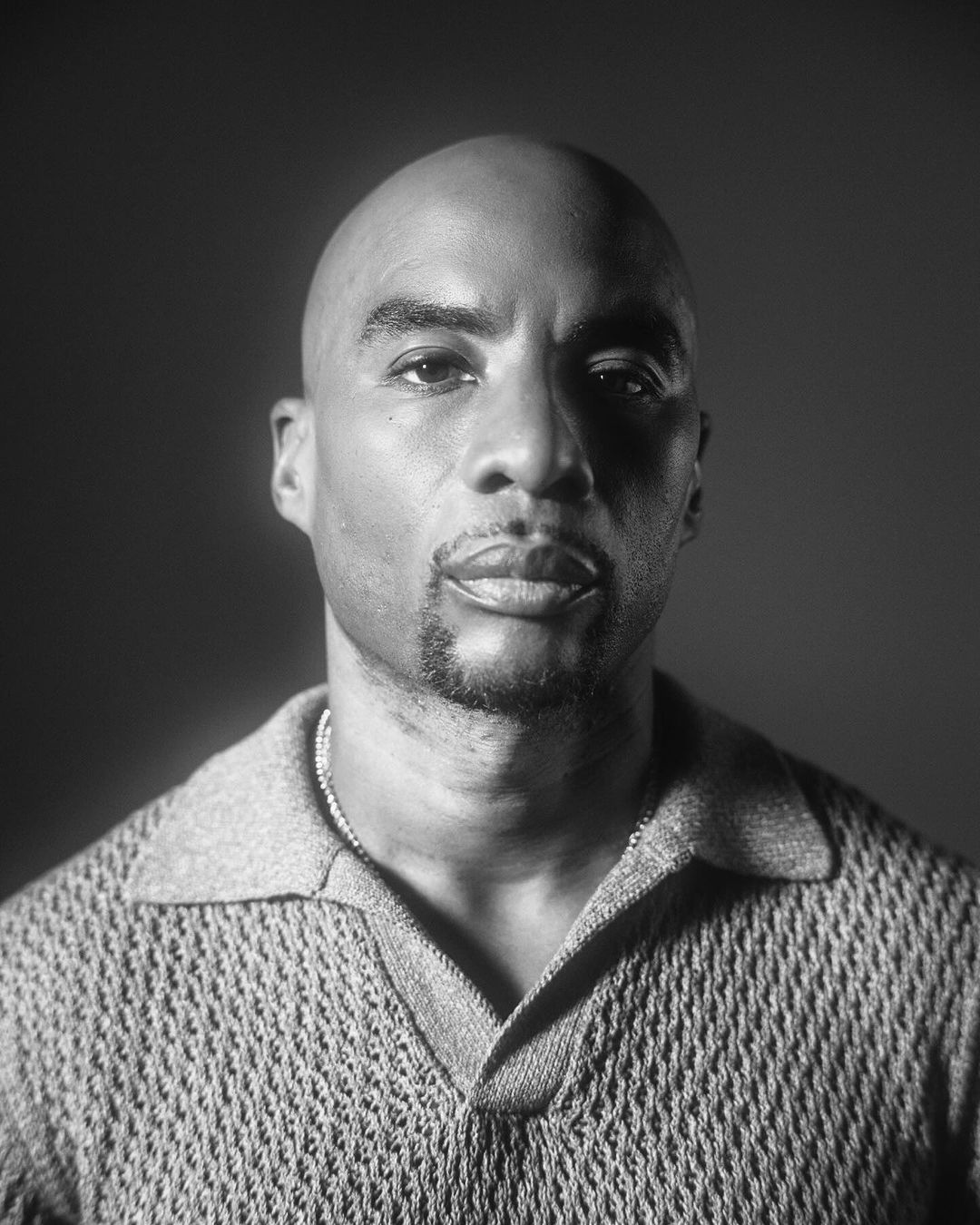 Monochrome portrait of a Black adult male with a bald head, wearing a collared shirt and chain, against a dark, neutral backdrop.