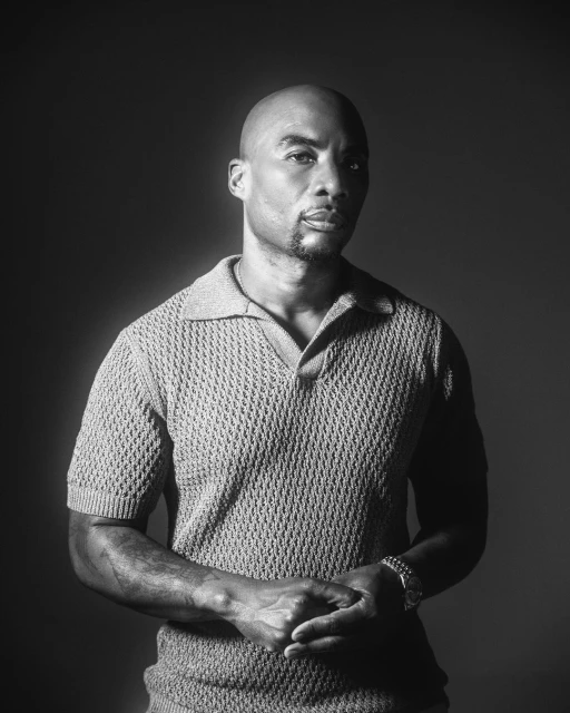 Monochrome portrait of a bald adult man in a short-sleeved polo shirt, gazing forward with folded hands and a wrist watch.
