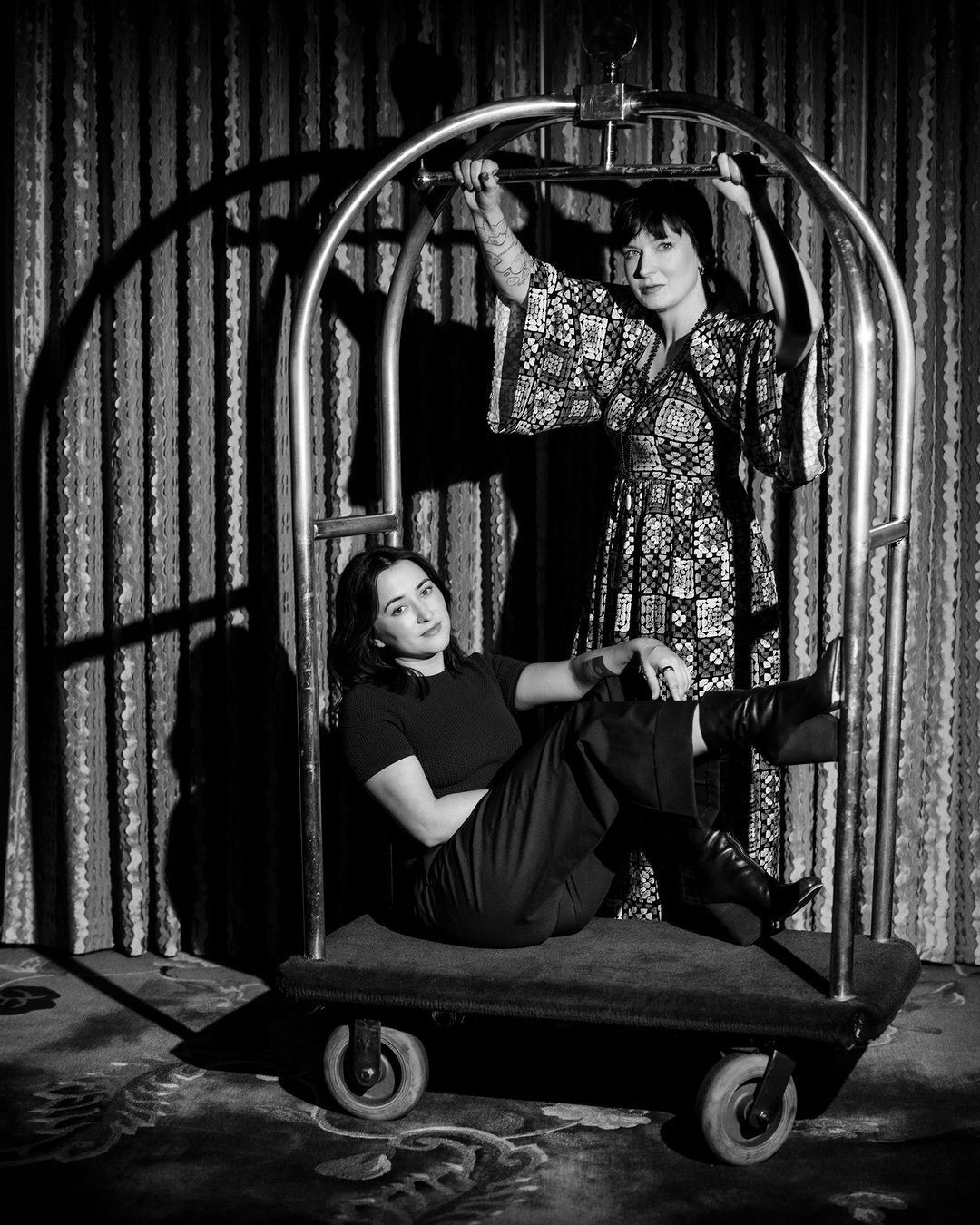 Two women pose inside luggage cart in black and white. One woman sits inside, another holds the cart. Patterned wall background.