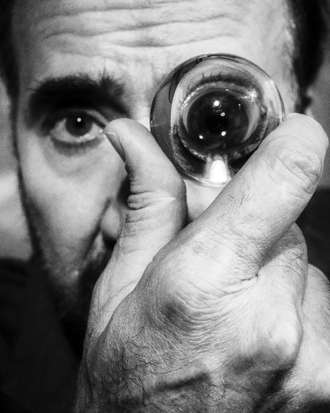 Black and white portrait of a man holding a clear crystal ball up to his eye, showcasing its refractive properties and the man's face.