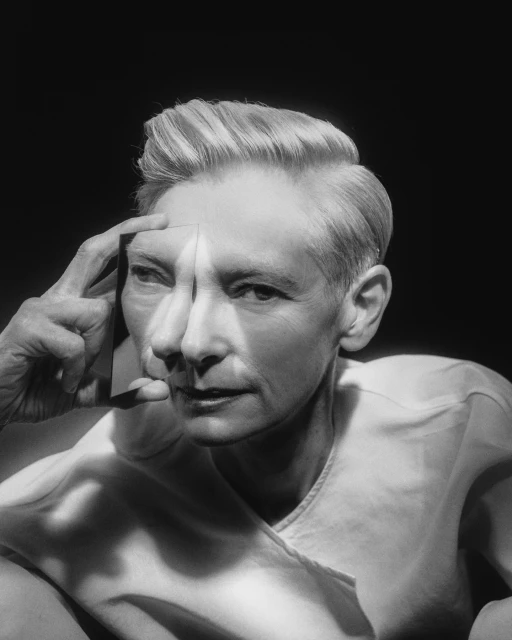 Monochromatic portrait of an adult with light hair with hand casting a shadow on their face in a high-contrast lighting studio setting.