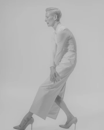 Monochrome full-length portrait of a person with short blonde hair wearing a tailored suit and heels against a white background.