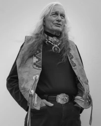 Portrait of an elderly man with long grey hair wearing a patterned leather vest and layered pendant necklace.