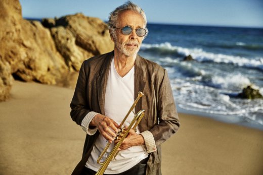 Elderly man with grey hair and sunglasses stands on a beach holding a trumpet with ocean, waves, and cliffs in the background.