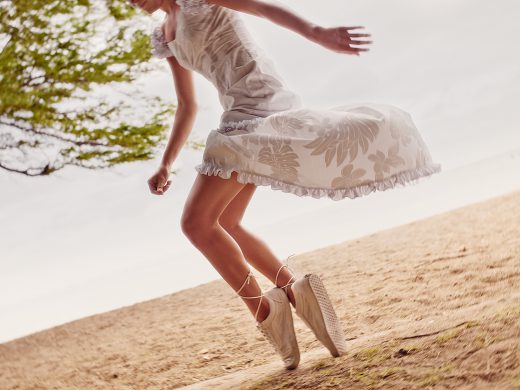 Woman in a white dress jumps on a sandy beach near a tree. The upward angle captures her movement, dress details, and the beach texture.